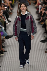 Model walking a fashion show in a gray and maroon color-block windbreaker over a navy sweater and white-collared shirt, paired with navy pants and white-blue sneakers.
