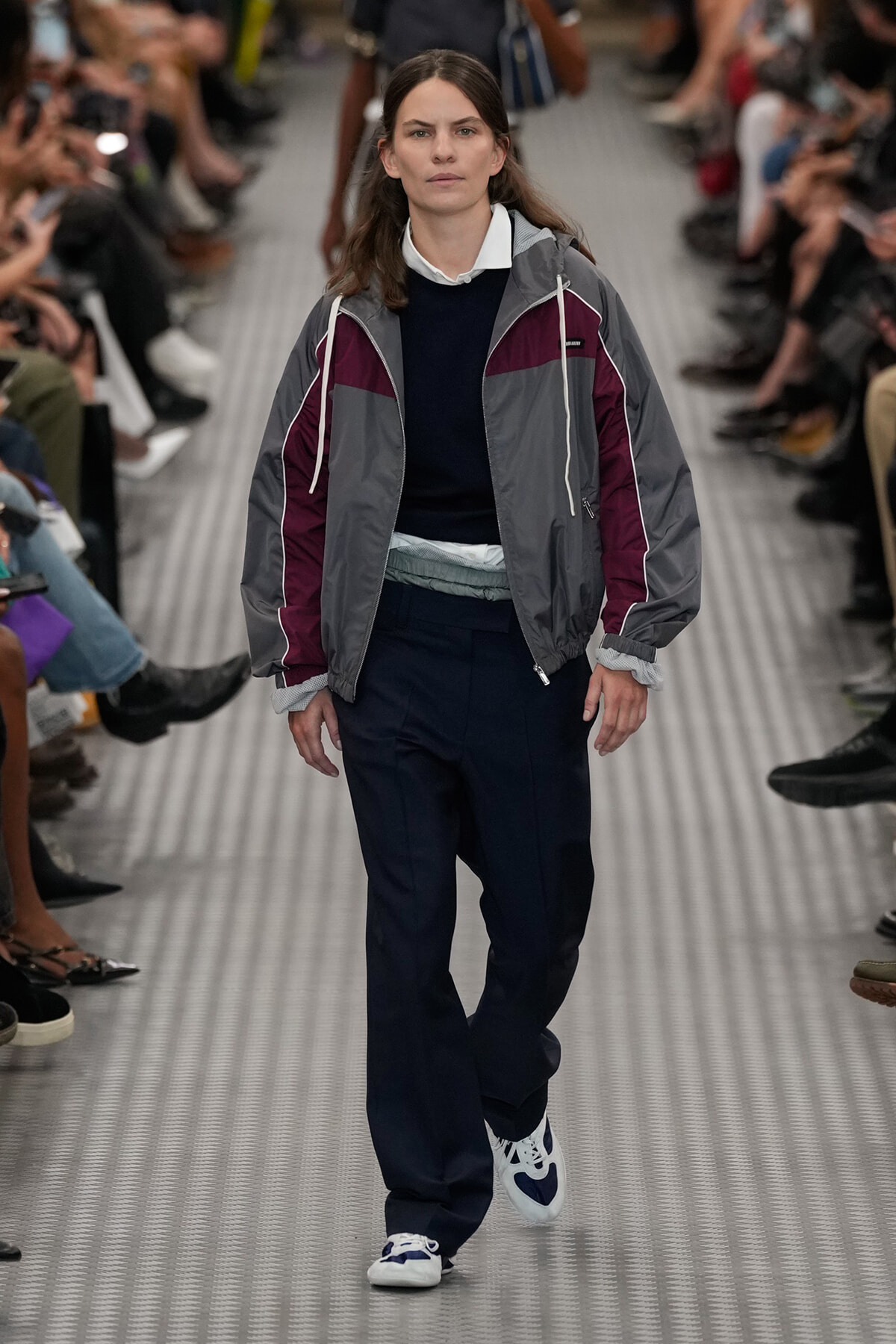 Model walking a fashion show in a gray and maroon color-block windbreaker over a navy sweater and white-collared shirt, paired with navy pants and white-blue sneakers.