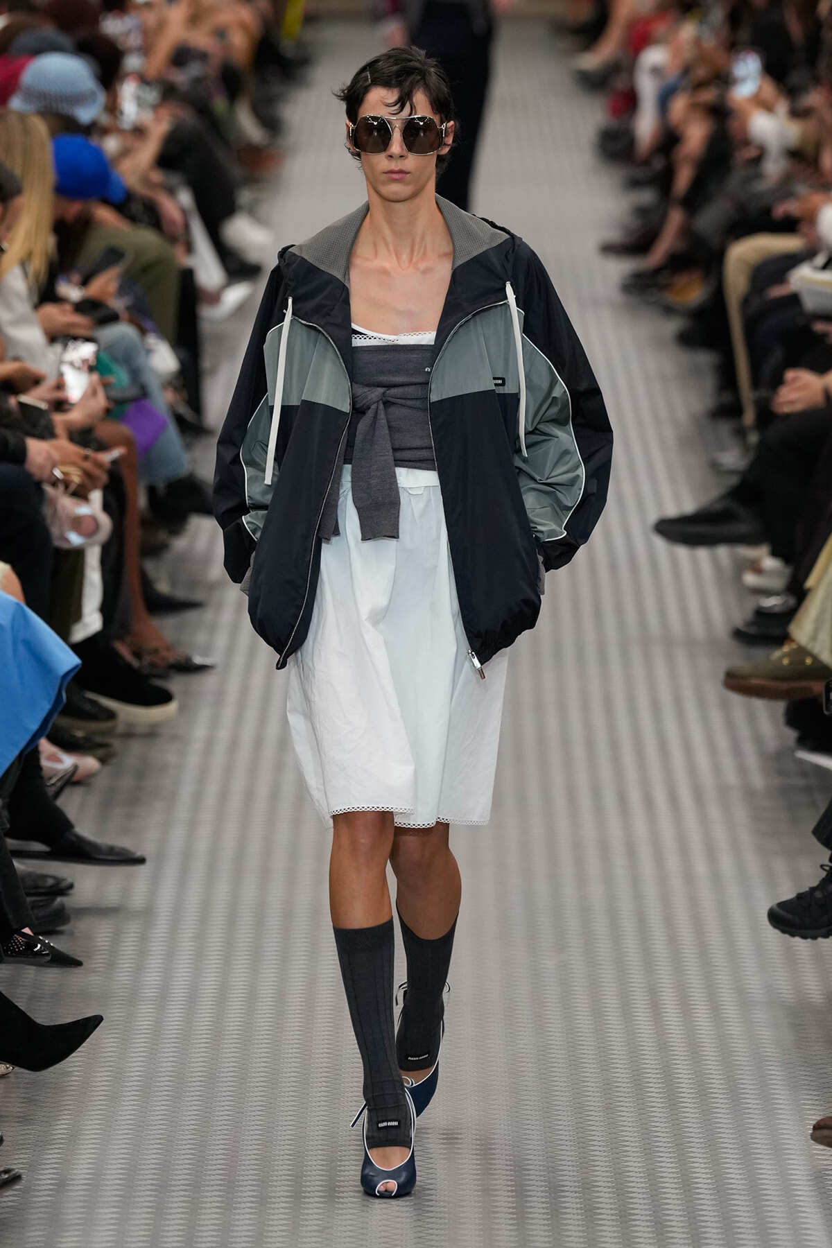 Model walks a runway in a color-block windbreaker over white shorts, knee-high socks, and navy open-toe shoes with oversized sunglasses.
