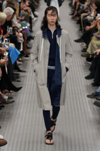 Model walking a fashion runway in a light gray long coat over navy layers, with a navy zip-up jacket and black sandals, audience seated on both sides.