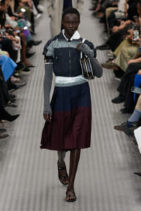 Runway model wearing a color-block windbreaker over a layered top with a navy and burgundy long skirt, gray arm warmers, sandals, and a black shoulder bag.
