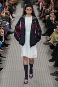 Model walking a runway in a white dress with a dark color-block windbreaker; audience seated along the sides.