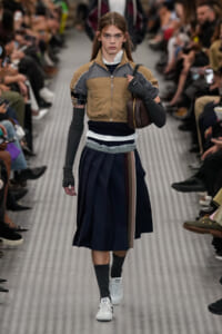 Model on a runway wearing a tan cropped windbreaker, navy pleated skirt, gray arm-warmers, white sneakers, and a brown shoulder bag.