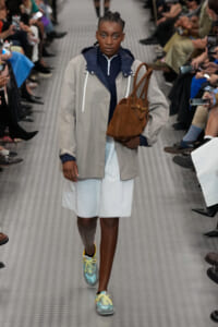 Model walking a fashion runway in a light gray rain jacket over a navy zip-top, white skirt, teal sneakers with yellow laces, and a brown tote bag over the shoulder.