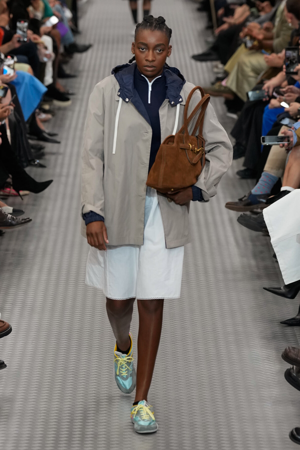 Model walking a fashion runway in a light gray rain jacket over a navy zip-top, white skirt, teal sneakers with yellow laces, and a brown tote bag over the shoulder.