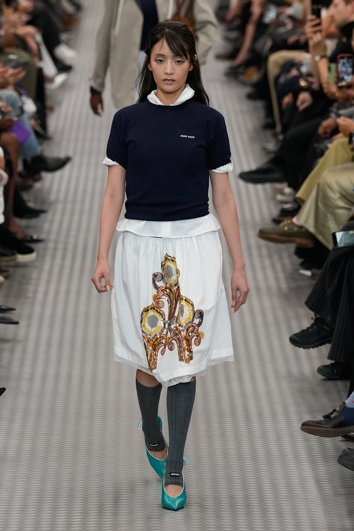 Model walking a fashion runway in a navy top, white layered skirt with a gold ornate motif, gray knee socks, and turquoise heels.