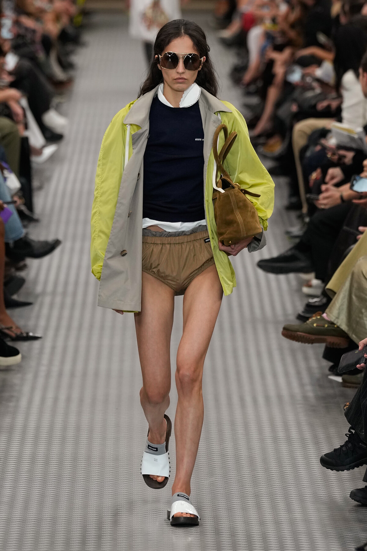 Model walks a runway in a lime-yellow and gray coat over a navy top, beige shorts, white slide sandals, oversized sunglasses, and a brown handbag.