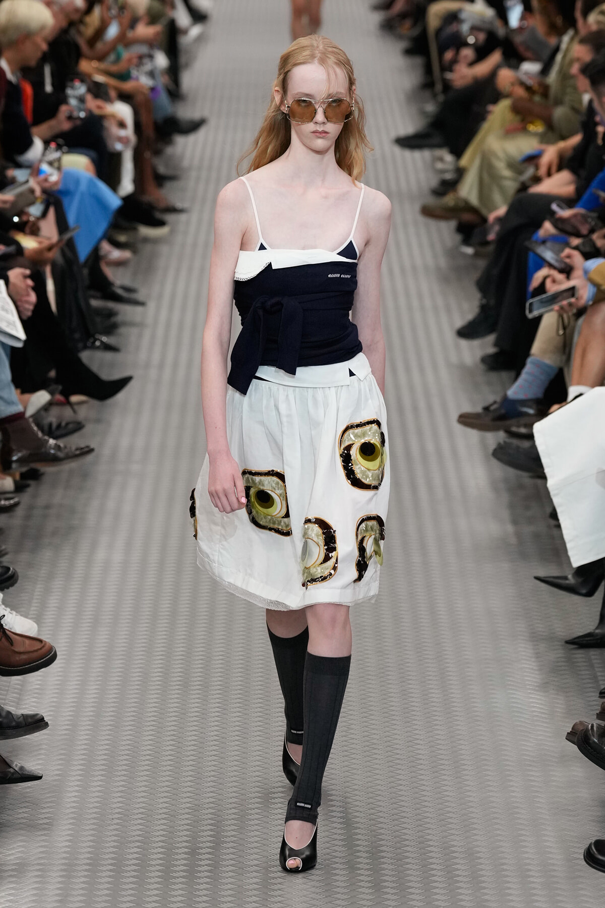 Model walks the runway in a two-tone navy and white dress with a belt, teamed with a white skirt featuring large eye motifs and black knee-high socks, at a fashion show.