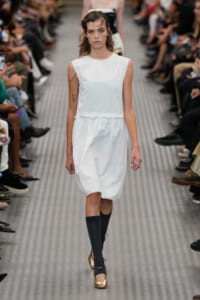 Model walking a fashion runway in a white sleeveless dress with an elastic waist, black knee-high socks, and gold peep-toe heels; audience sits along the sides.
