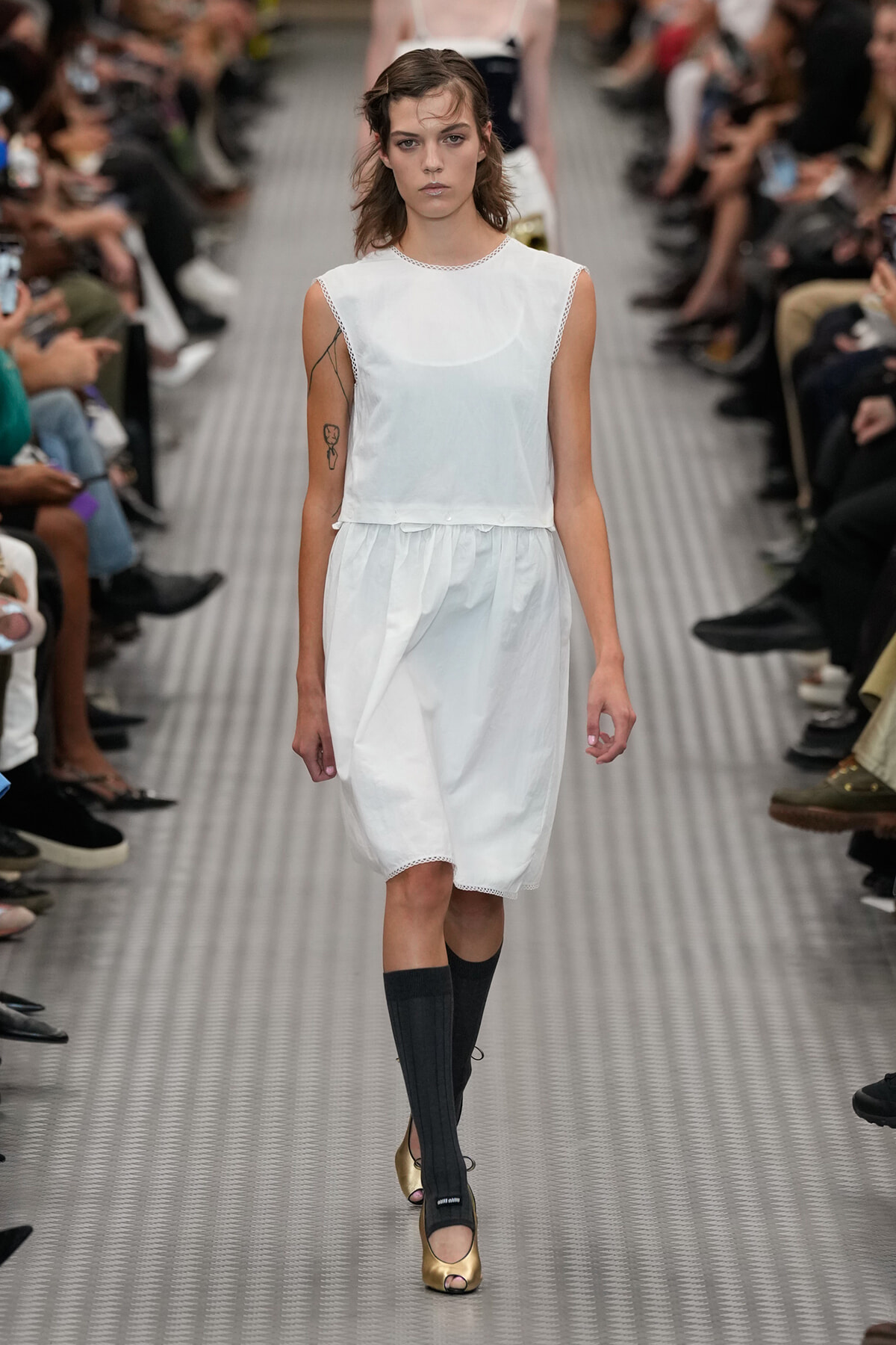 Model walking a fashion runway in a white sleeveless dress with an elastic waist, black knee-high socks, and gold peep-toe heels; audience sits along the sides.
