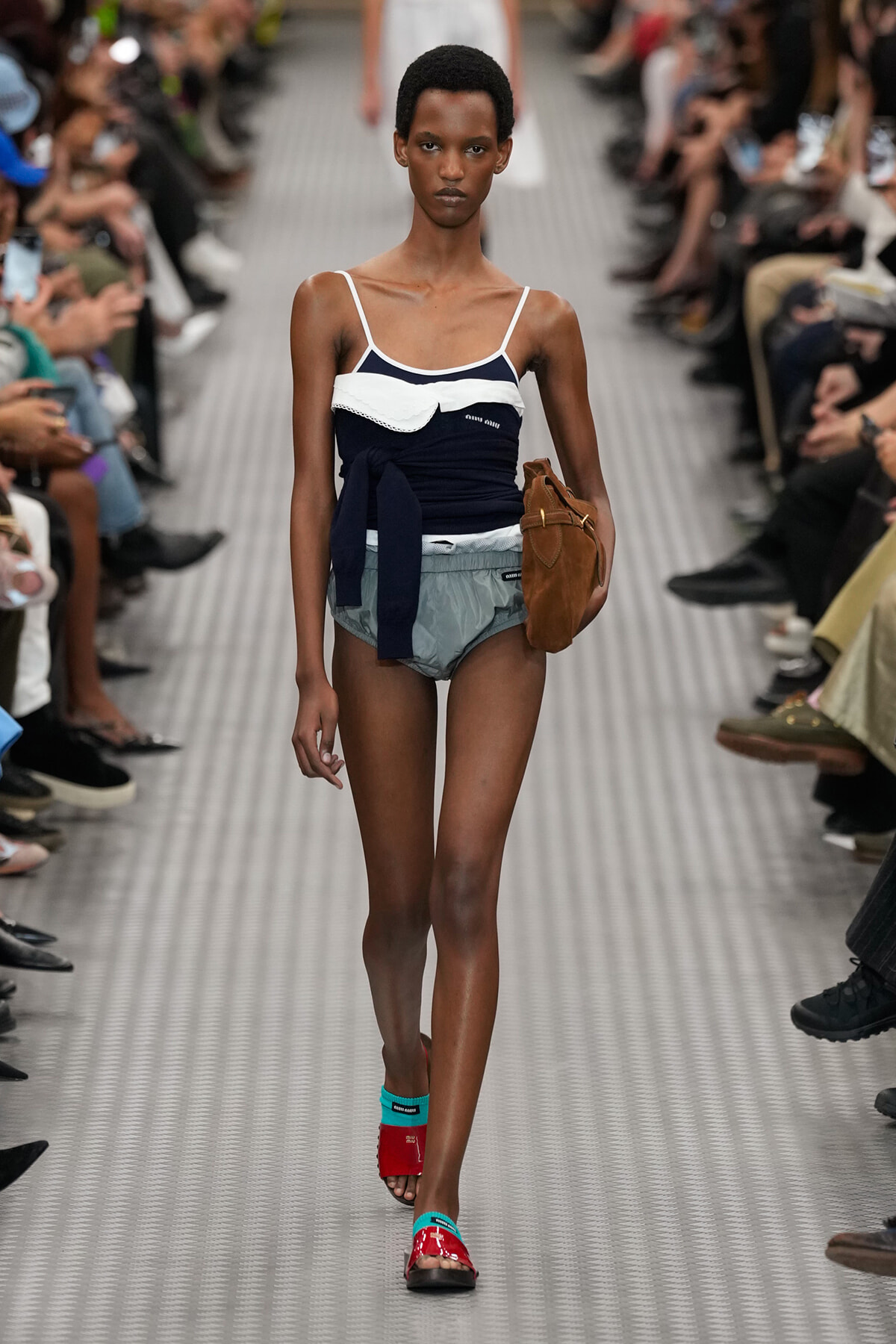 Model walks a fashion runway in a navy top with white trim, light gray shorts, brown handbag, and red sandals with turquoise accents.