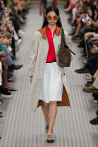 Model walks a runway wearing a beige trench with brown collar over a red top and light-blue skirt, carrying a brown tote bag and wearing oversized sunglasses.
