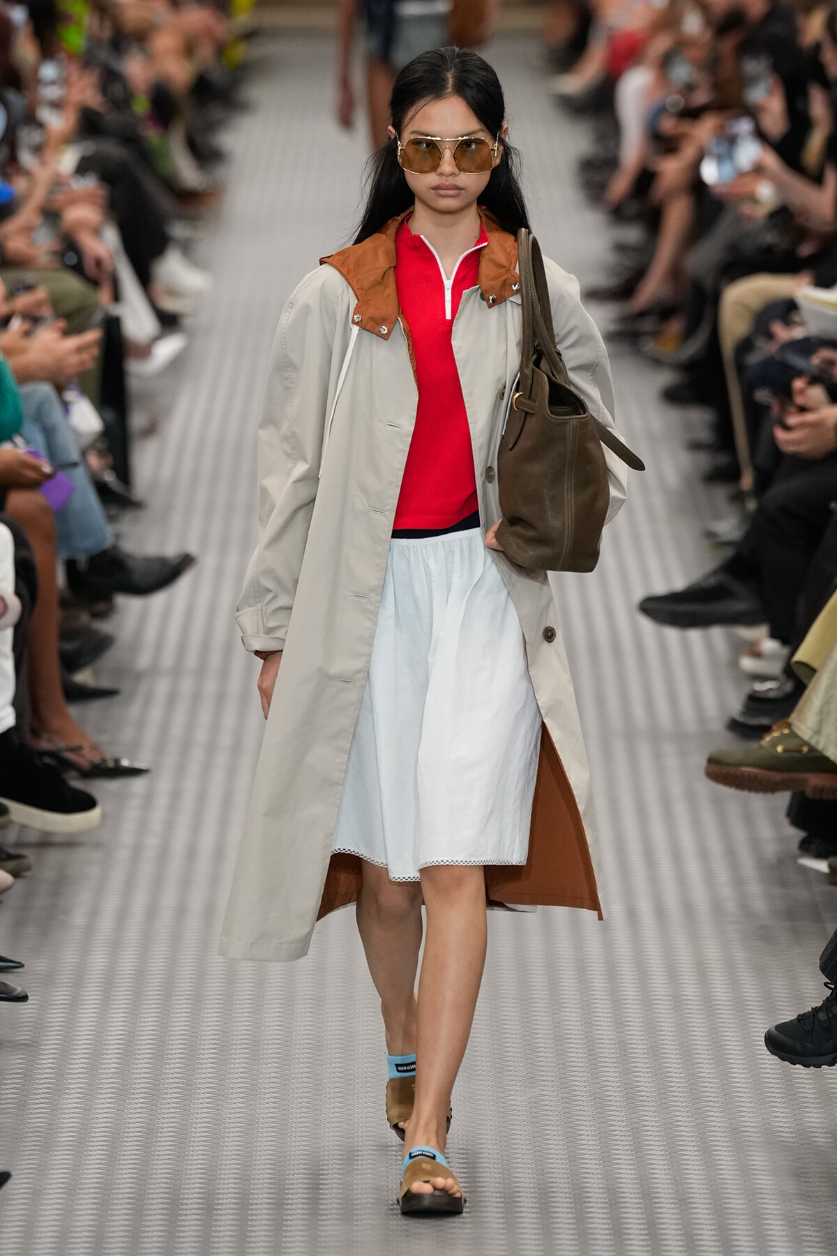 Model walks a runway wearing a beige trench with brown collar over a red top and light-blue skirt, carrying a brown tote bag and wearing oversized sunglasses.