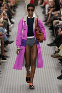 Model walking a runway in a pink-purple trench over a navy top and gray shorts, carrying a brown handbag and wearing oversized sunglasses.