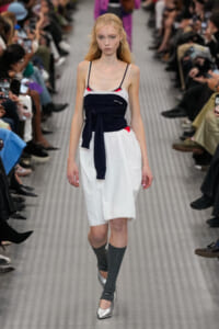 Model walking a fashion runway in a navy wrap top over a white A-line skirt, gray knee-high socks, and silver flats, audience on both sides