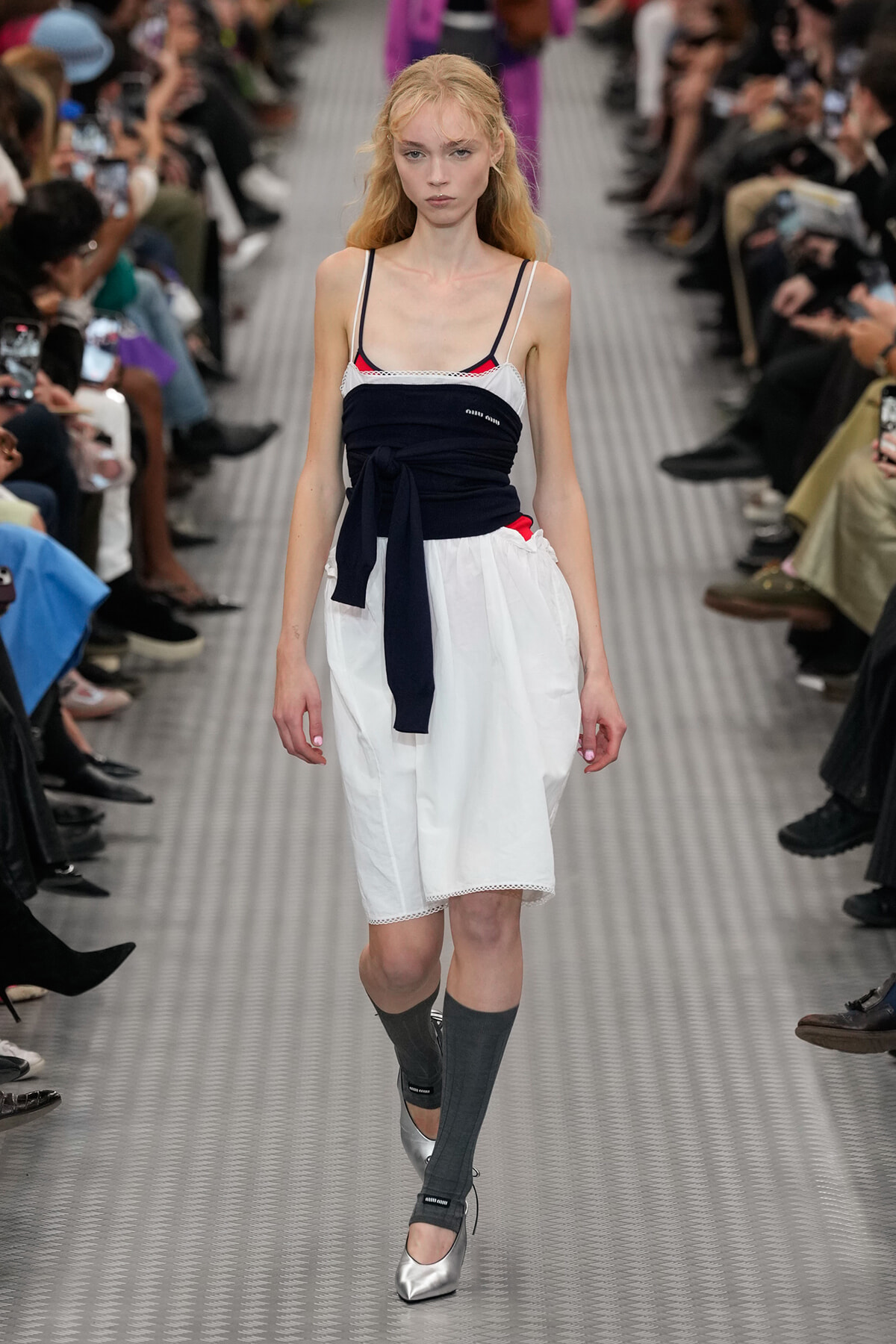 Model walking a fashion runway in a navy wrap top over a white A-line skirt, gray knee-high socks, and silver flats, audience on both sides