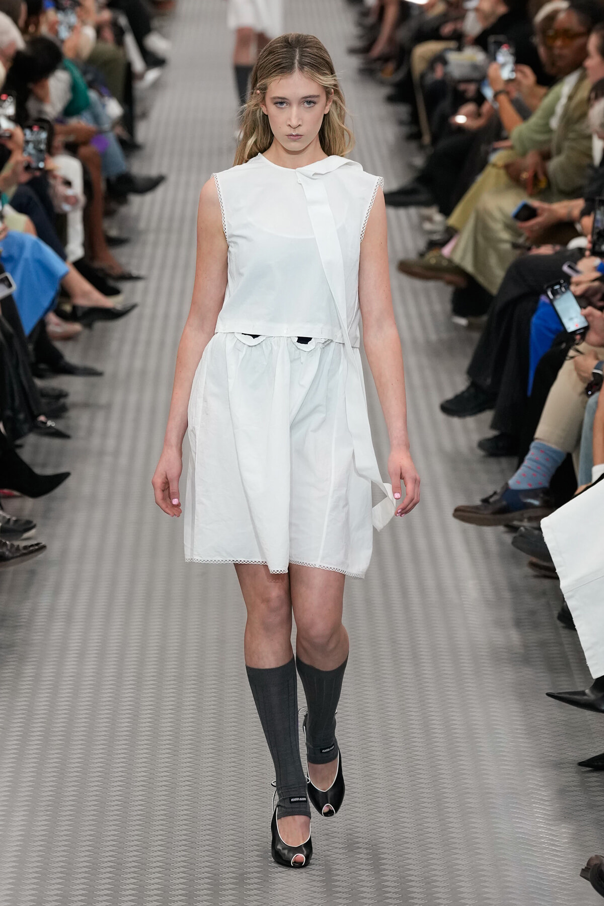 Model walking the runway in a white sleeveless dress with a wrap detail and knee-length skirt, paired with gray knee-high socks and black open-toe heels.