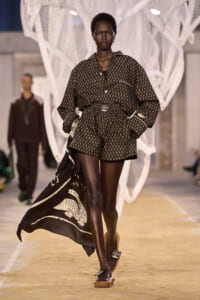 Black female model walks a fashion runway in a brown patterned shirt and shorts with a belted waist and flowing cape behind her on a beige catwalk.