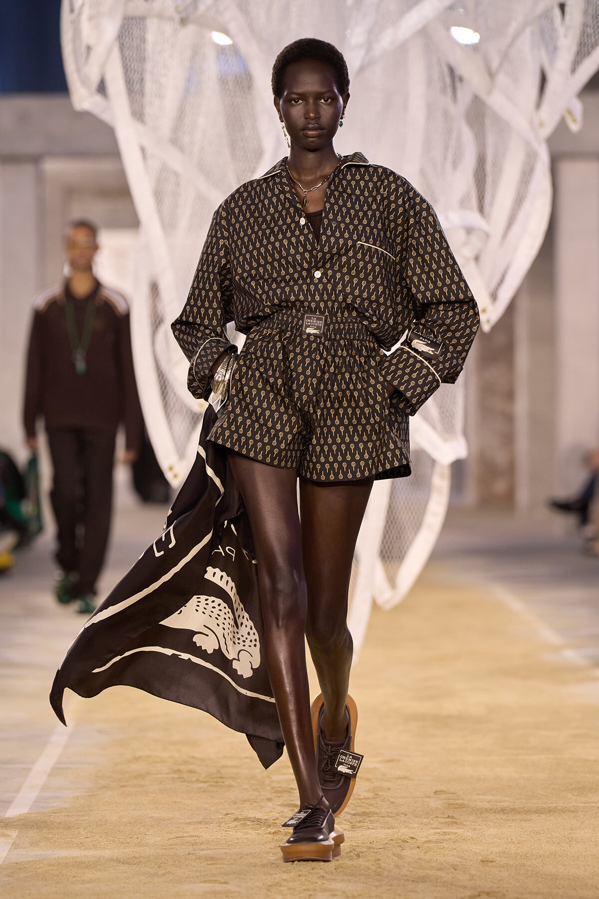 Black female model walks a fashion runway in a brown patterned shirt and shorts with a belted waist and flowing cape behind her on a beige catwalk.