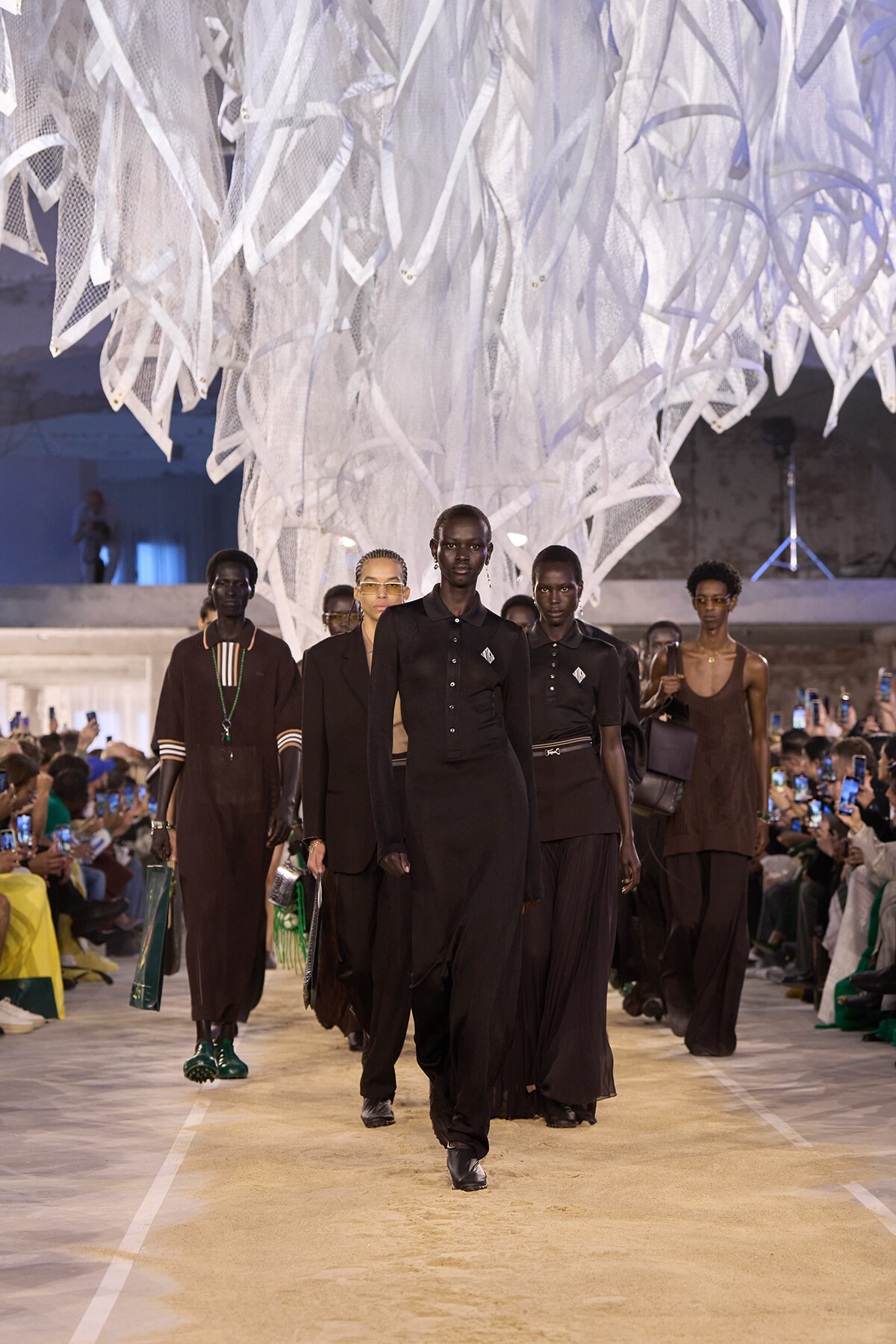 Models walk a runway under hanging white fabric sculptures as spectators film from the sides.