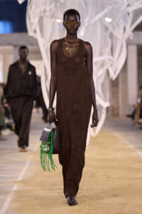 Model with short hair walks a fashion runway in a brown crochet maxi dress, accessorized with green jewelry and a black bag with neon green fringe.