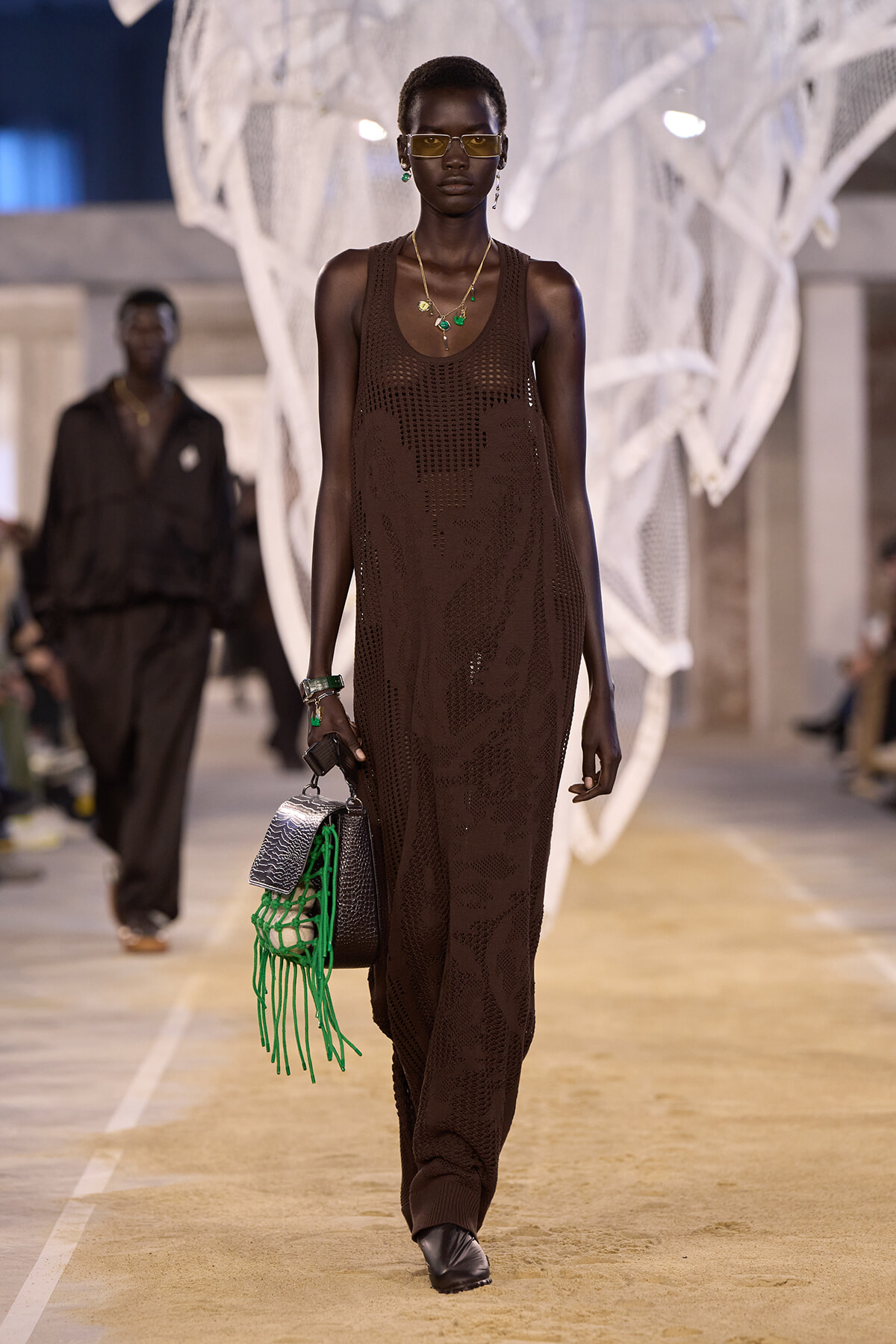 Model with short hair walks a fashion runway in a brown crochet maxi dress, accessorized with green jewelry and a black bag with neon green fringe.