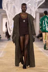 Model walking the runway in a dark brown bodysuit under a long patterned cape with a geometric print and green jewelry.