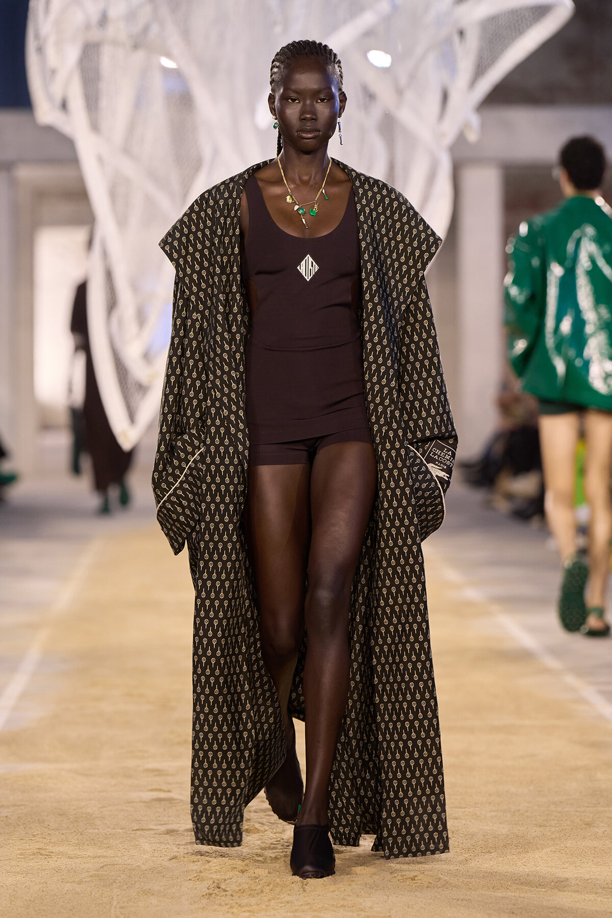 Model walking the runway in a dark brown bodysuit under a long patterned cape with a geometric print and green jewelry.