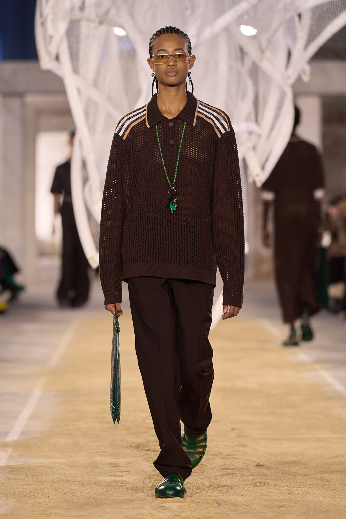 Model walking a runway in a brown mesh polo sweater, dark trousers, green sandals, and yellow-tinted glasses, holding a green bag.