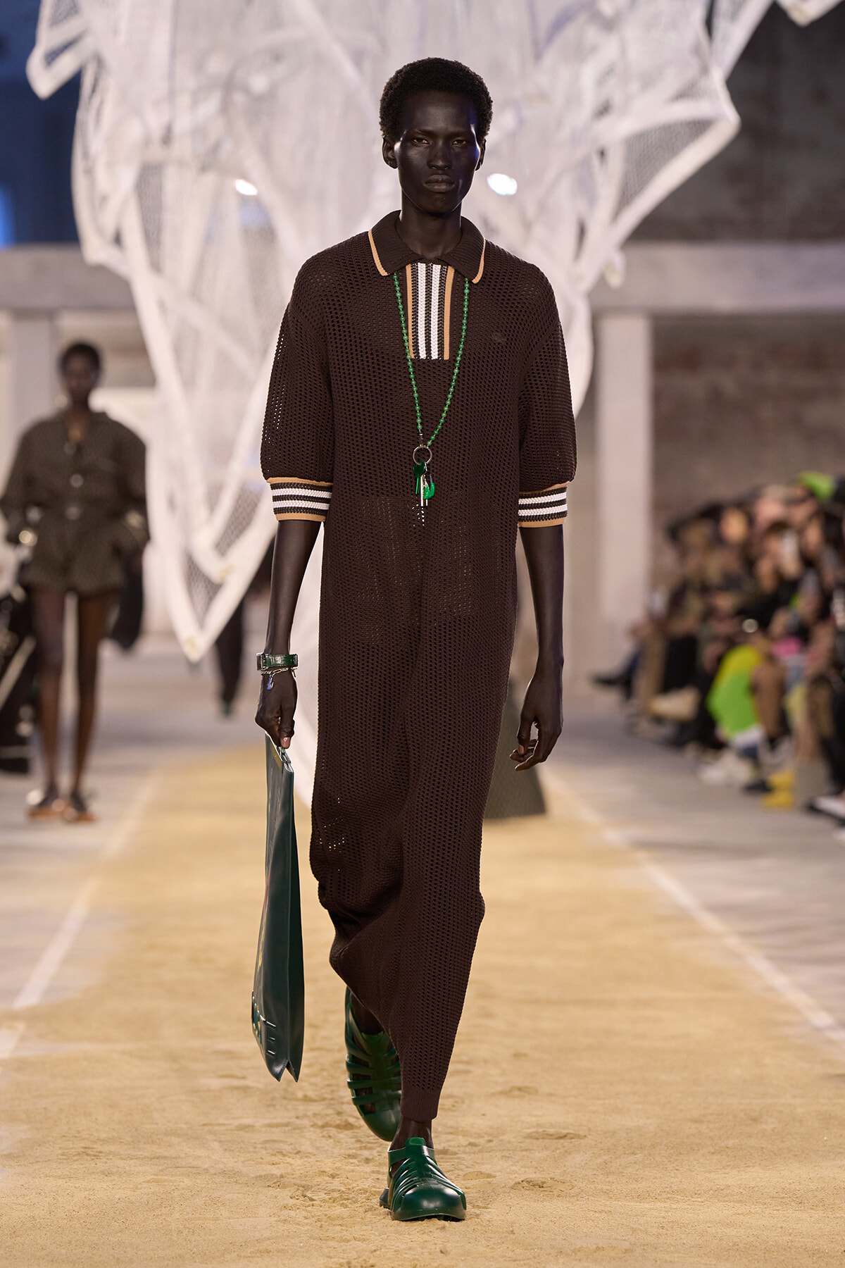 Male model walking a fashion runway in a brown knit long-sleeve dress with striped collar and cuffs, accessorized with green jewelry and a green geometric bag.