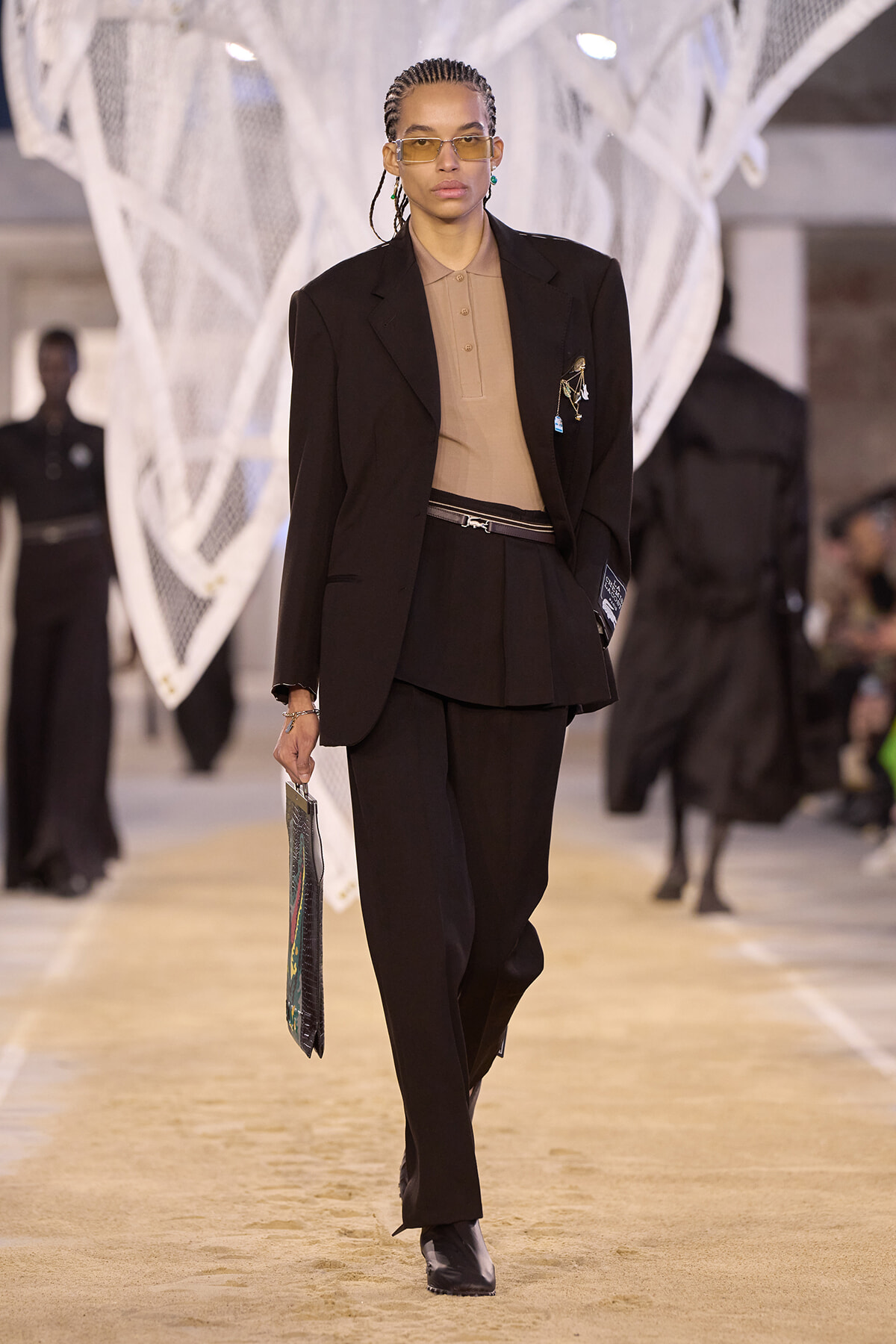 Model walks a fashion runway in a black suit with a tan polo top, holding a patterned clutch.