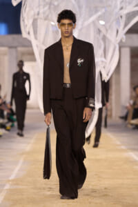 Male model walking a fashion runway in a black suit with a tan top, wearing tinted glasses and carrying a black portfolio.
