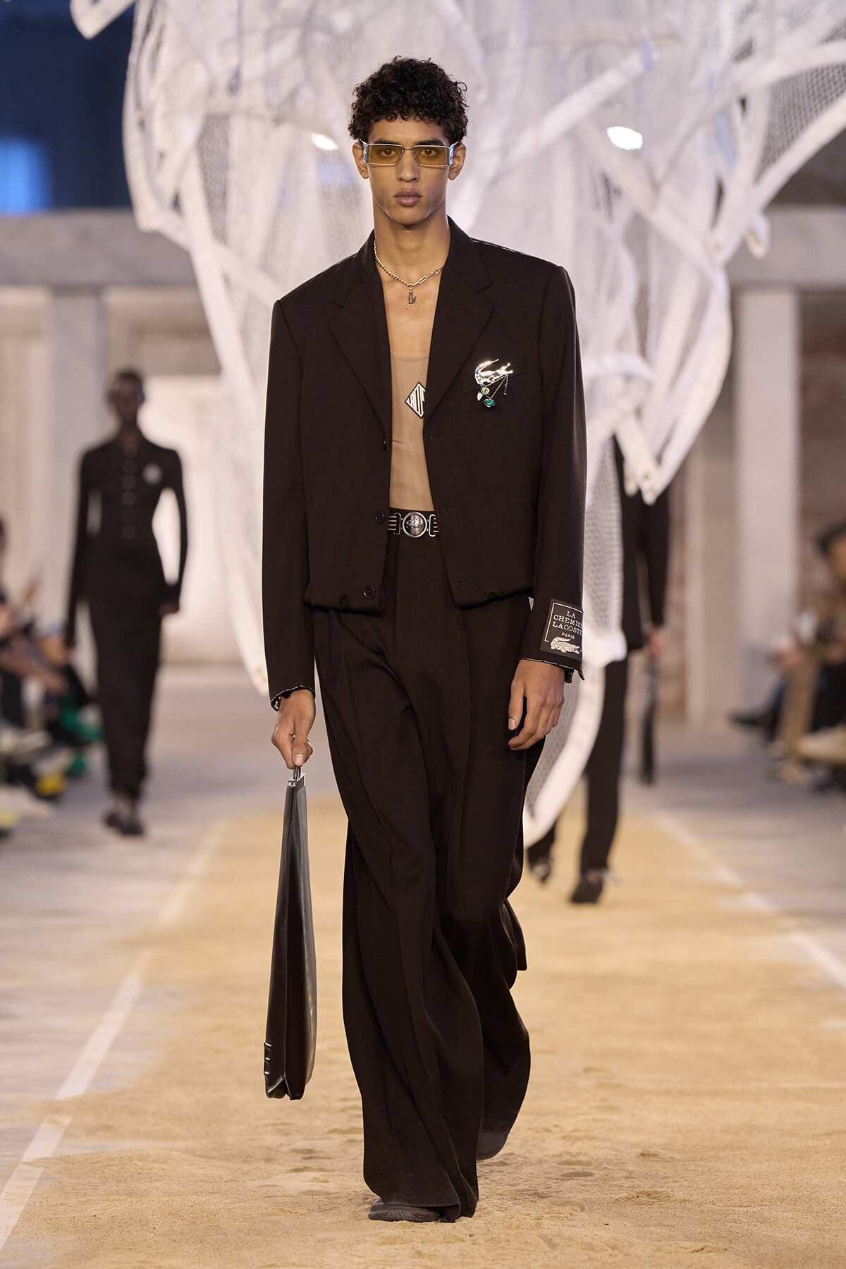 Male model walking a fashion runway in a black suit with a tan top, wearing tinted glasses and carrying a black portfolio.