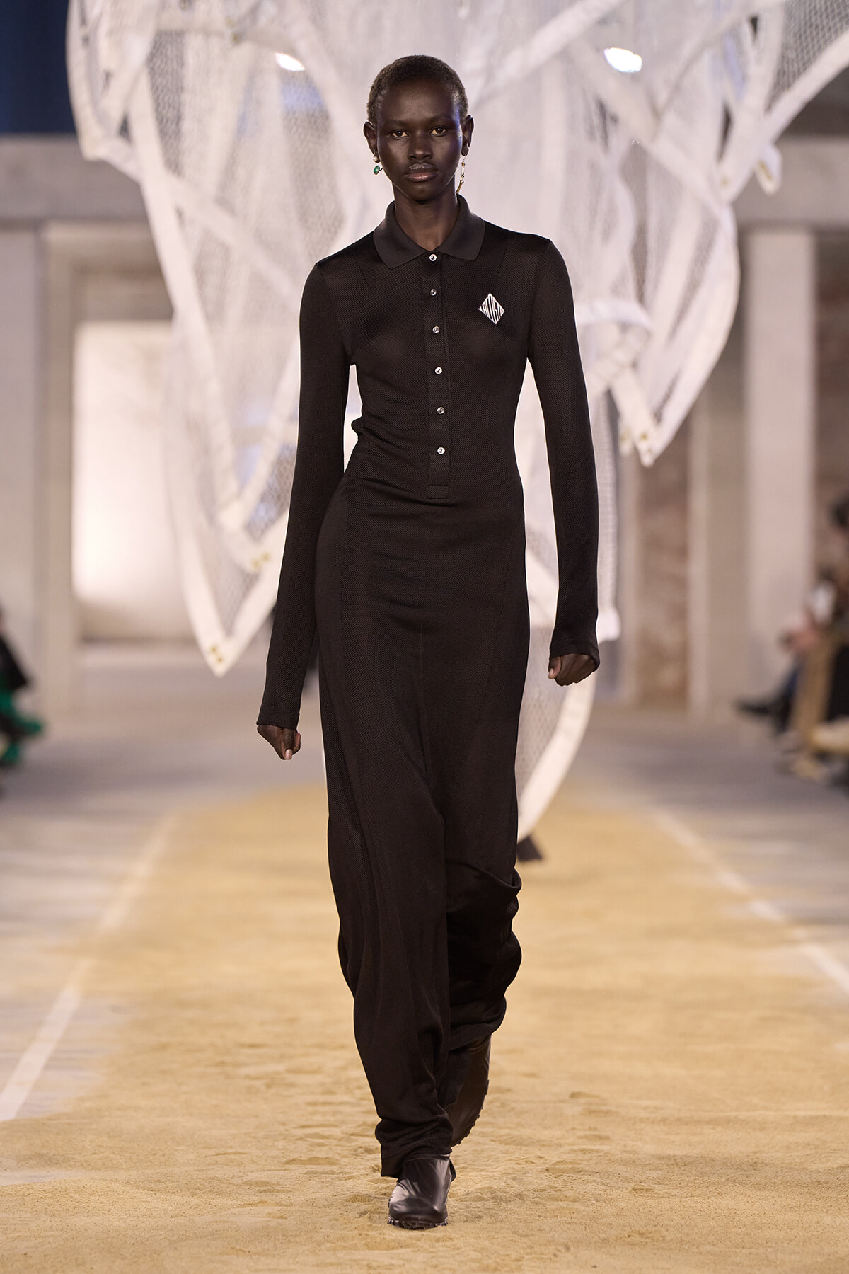 Model walking the runway in a black long-sleeve knit polo jumpsuit with a button placket and small chest logo, against a white sculptural backdrop.