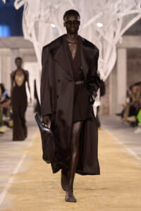 Runway model in an oversized black coat with a deep V, black shorts, sunglasses, and a textured black clutch on a beige catwalk.