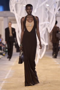Model walking a fashion runway in a long dark brown knit gown over a beige top, carrying a small handbag.