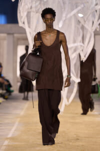Male runway model wearing a brown sleeveless knit top with wide brown pants, carrying a large dark leather bag on the runway.
