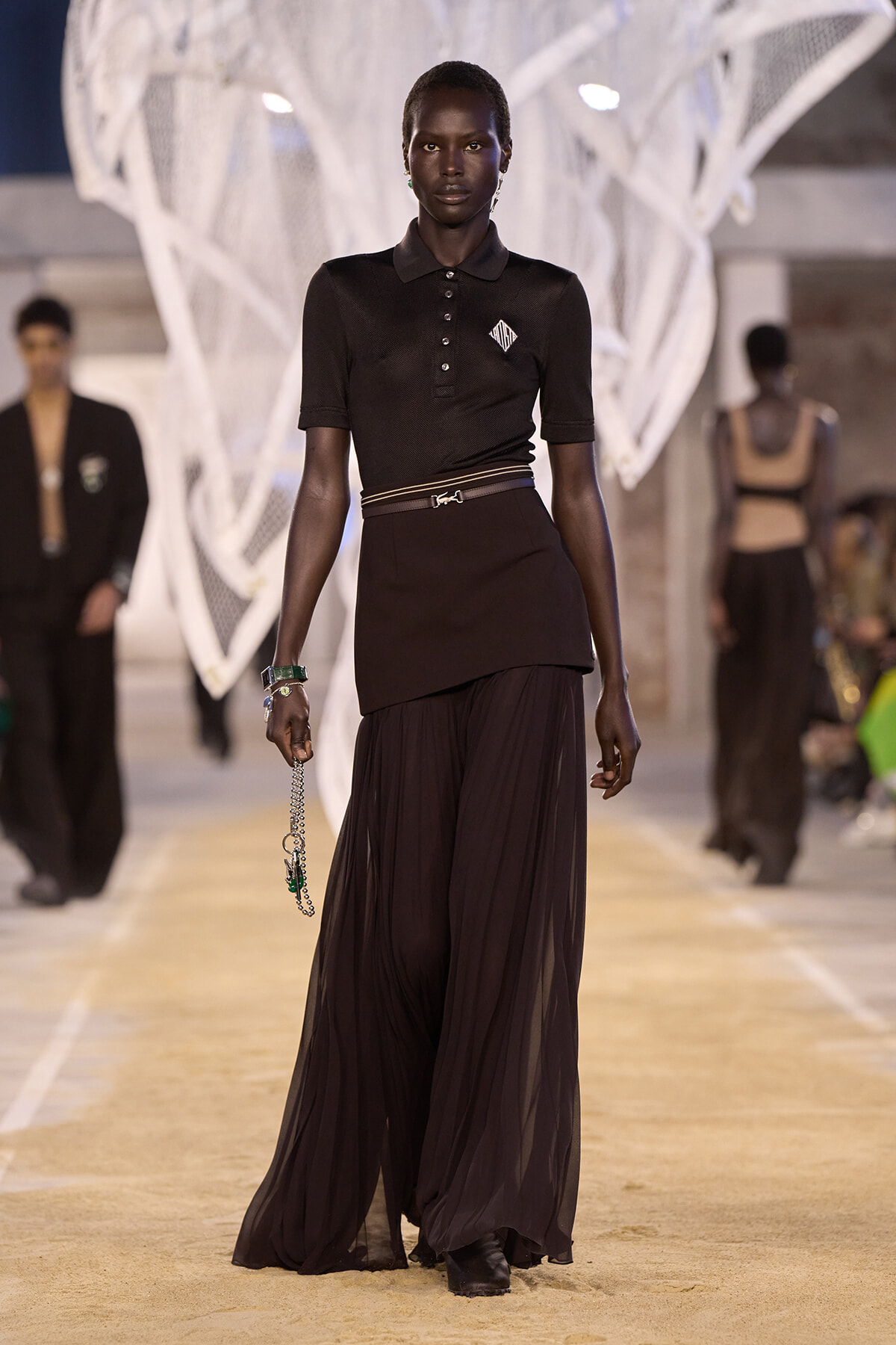 Model walking a runway in a black polo top and flowing black skirt with a slim belt, holding a chain accessory.