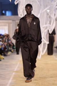 Male model walking a runway in a black, satin tracksuit with zipper and gathered cuffs, necklace visible below open chest.