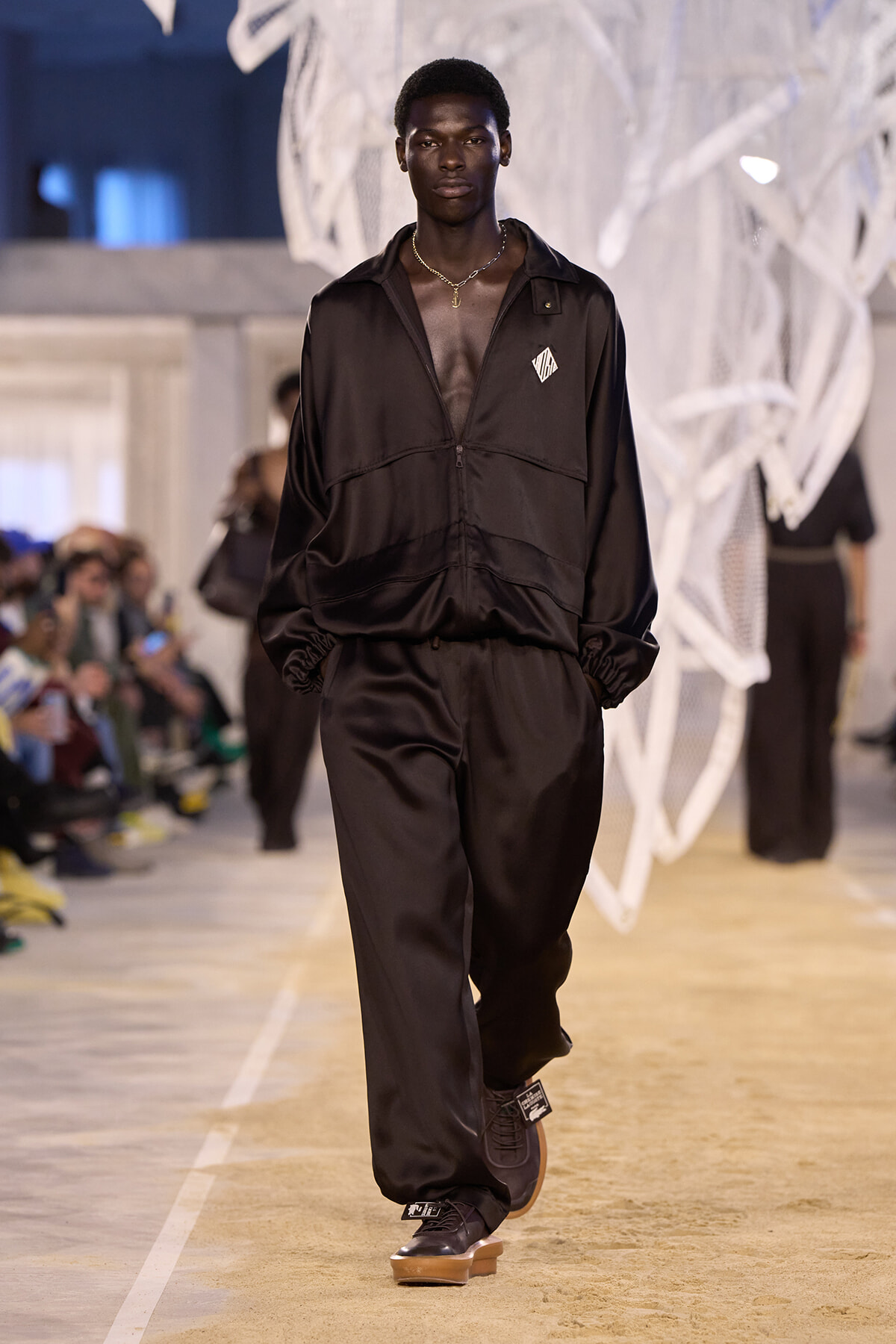 Male model walking a runway in a black, satin tracksuit with zipper and gathered cuffs, necklace visible below open chest.