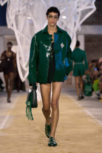 Male model on a fashion runway wearing a shiny dark green patent leather jacket, black attire, sunglasses, and a green fringe bag.