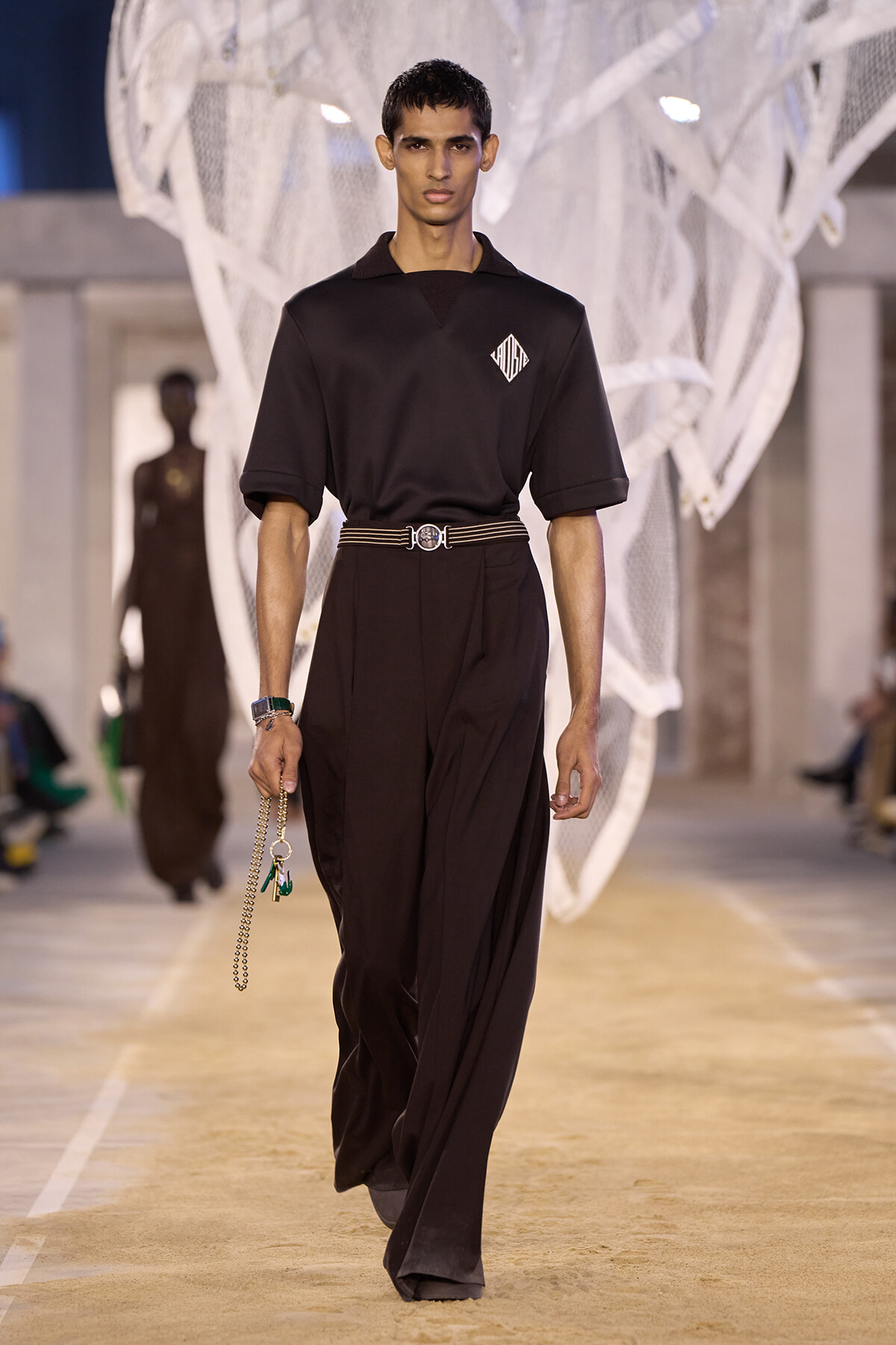 Male model walks a fashion runway in an all-black look: loose polo, wide dark trousers, striped belt, and a chain accessory dangling from his hand.