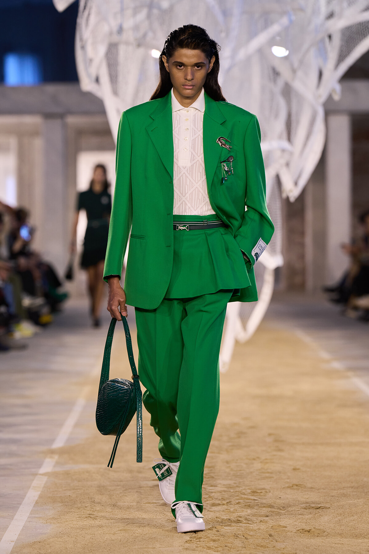 Model walking a fashion runway in a bright emerald green suit with a peplum detail, white knit top, and white sneakers; carrying a dark green textured handbag.