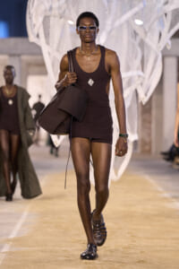 Person walking a fashion runway in a dark brown sleeveless top and matching shorts, carrying a large brown bag and wearing sunglasses and a necklace.
