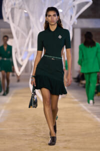 Model walking a green polo dress with a pleated hem on a fashion runway, carrying a patterned handbag.