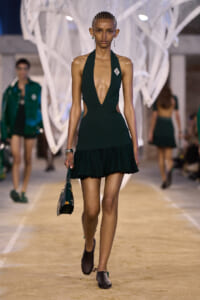 Model walks the runway in a dark emerald green halter dress with a deep plunging neckline and a flouncy mini hem, carrying a green handbag.