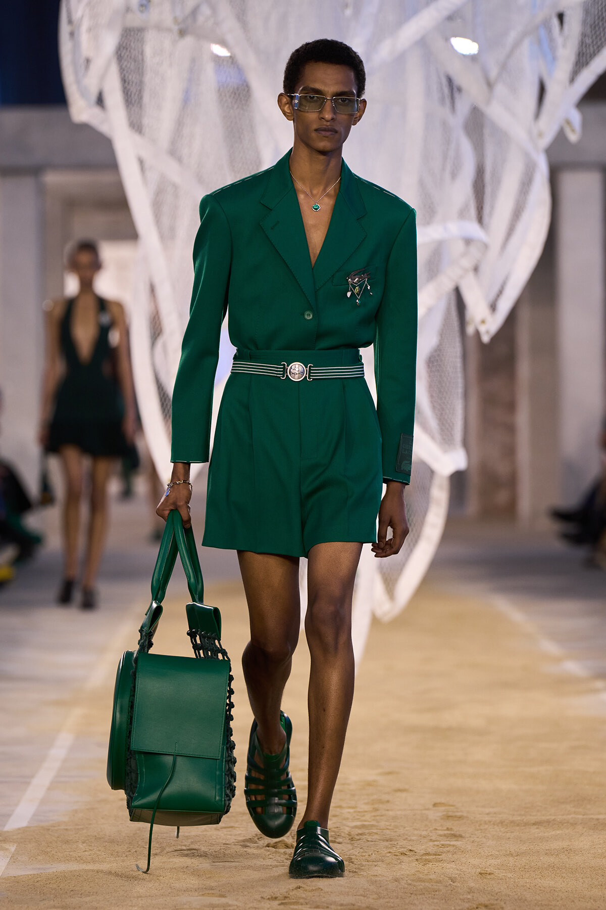 Model in a green blazer and matching shorts walking a runway, carrying a green handbag and wearing sunglasses and a necklace.