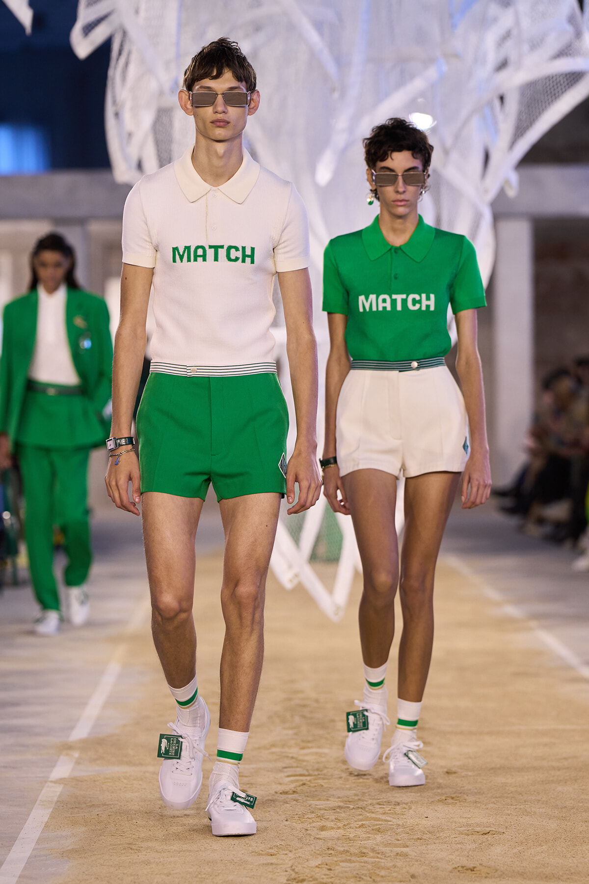 Two models walk a runway wearing MATCH-branded outfits: a white polo with green text and green shorts, plus white sneakers and sunglasses for both.