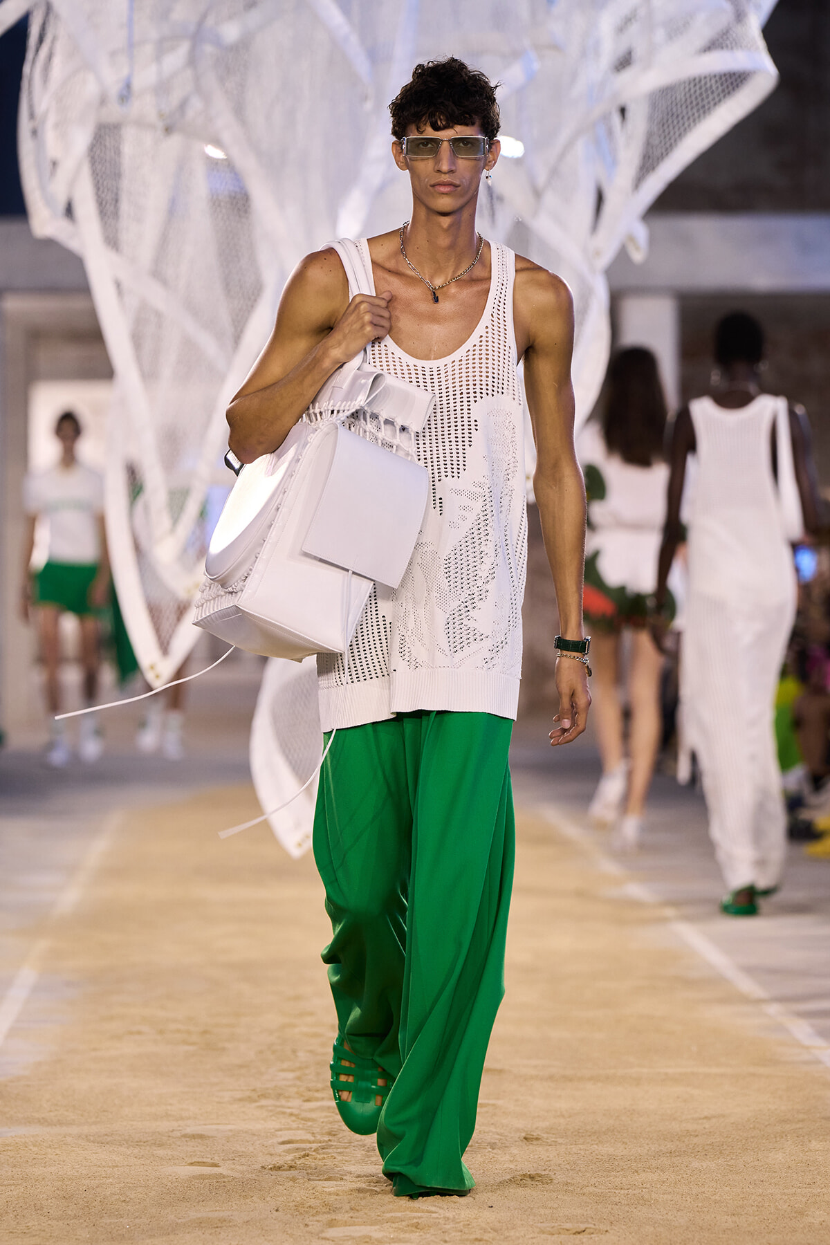 Male model on a runway wearing a white mesh tank, emerald green wide-leg pants, and a large white backpack with sunglasses.