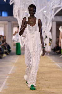 Model walks a runway in a white crochet maxi dress with a white shoulder bag and bright green shoes.